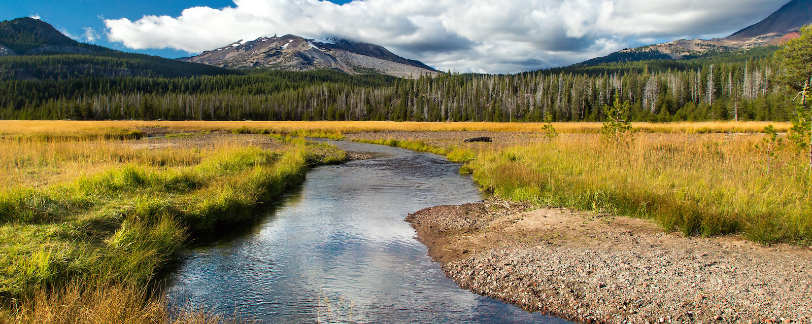 Stream and golden fields in foreground, mountains and clouds in background.